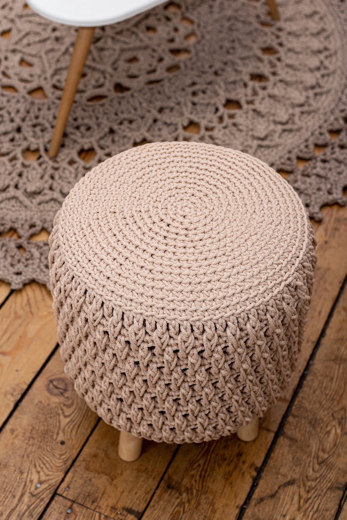 Heegeldatud tumba: Skandinaavia stiilis mugavus sinu elutoas A cozy crochet ottoman placed on a patterned rug and wooden floor, ideal for interior design inspiration.