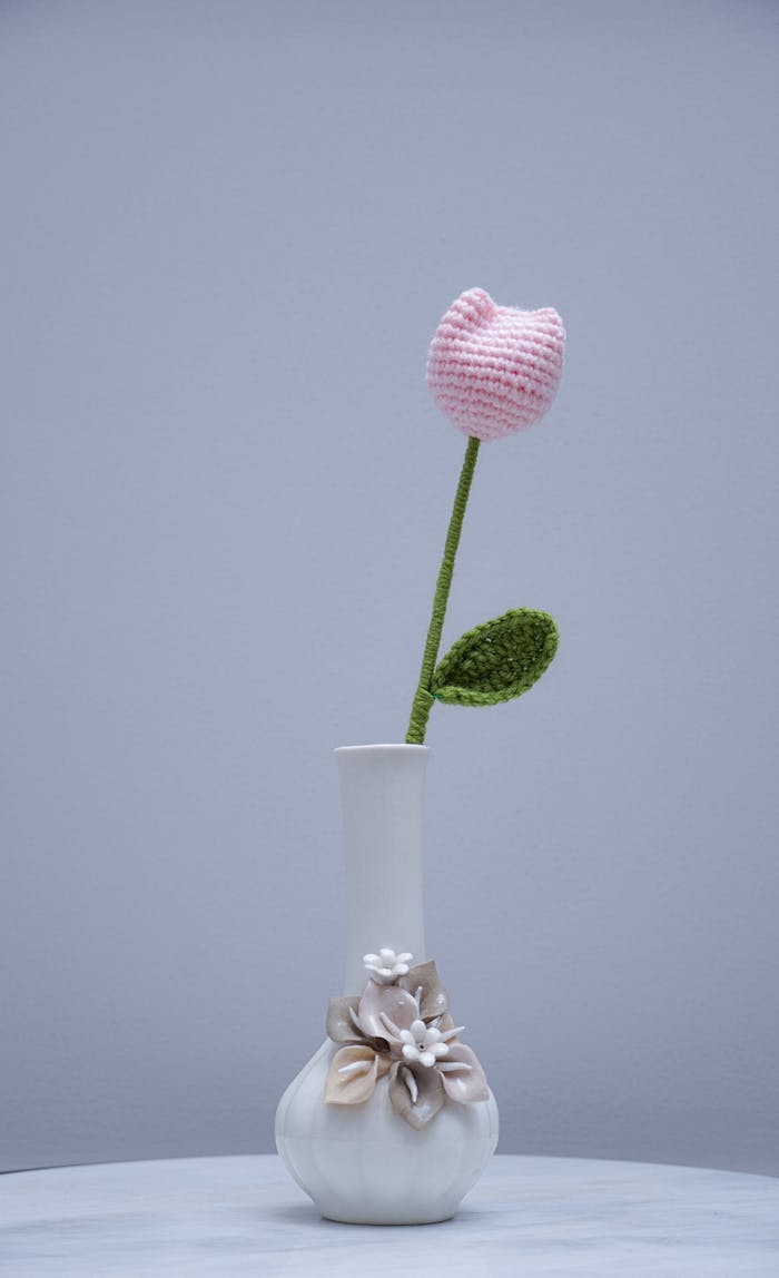 Igavesti õitsev ilu: Heegeldatud tulbid ja lilled – kingitus, mis kestab (+tasuta muster!) Pink crochet flower in artisanal ceramic vase with floral design on gray background.