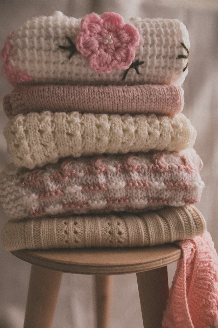 Hubane heegeldatud pleed: Miks see on sinu kodu peidetud pärl ja kuidas seda ise luua (tasuta muster!) A serene stack of soft knitted sweaters in pastel shades on a wooden stool, perfect for a cozy vibe.