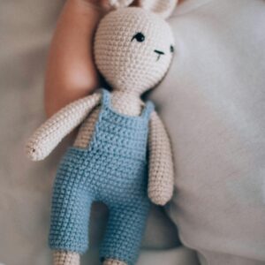 A cute baby lies next to a handmade crochet bunny doll in soft lighting.