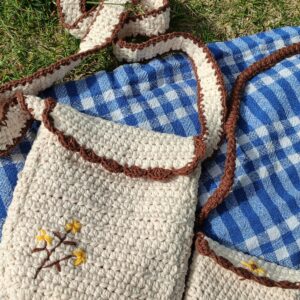 A crochet bag with floral embroidery rests on a checkered picnic blanket outdoors.